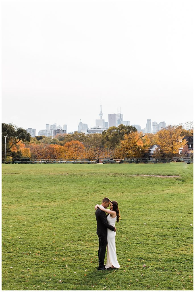 Bride and groom hug in field with city skyline and fall trees in background| Toronto wedding photographer| Toronto engagement photographer| 3photography 