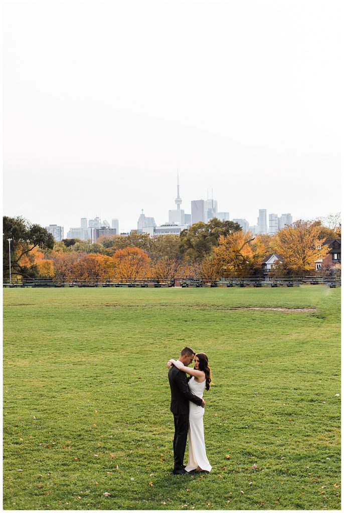 Bride and groom hug in field with city skyline and fall trees in background| Toronto wedding photographer| Toronto engagement photographer| 3photography